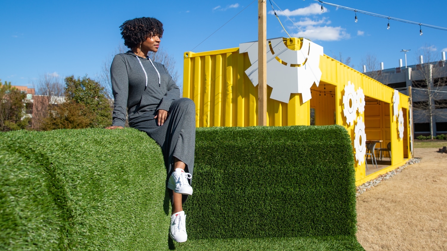 Delisia Matthews wears a long grey dress with with white drawstrings at the collar and long sleeves. She sits on top of a larger-than-life green couch made out of synthetic turf with her legs crossed and looking straight ahead but away from the camera, which is positioned to her right. Behind her, a remodeled shipping container that has been turned into a work space is visible.