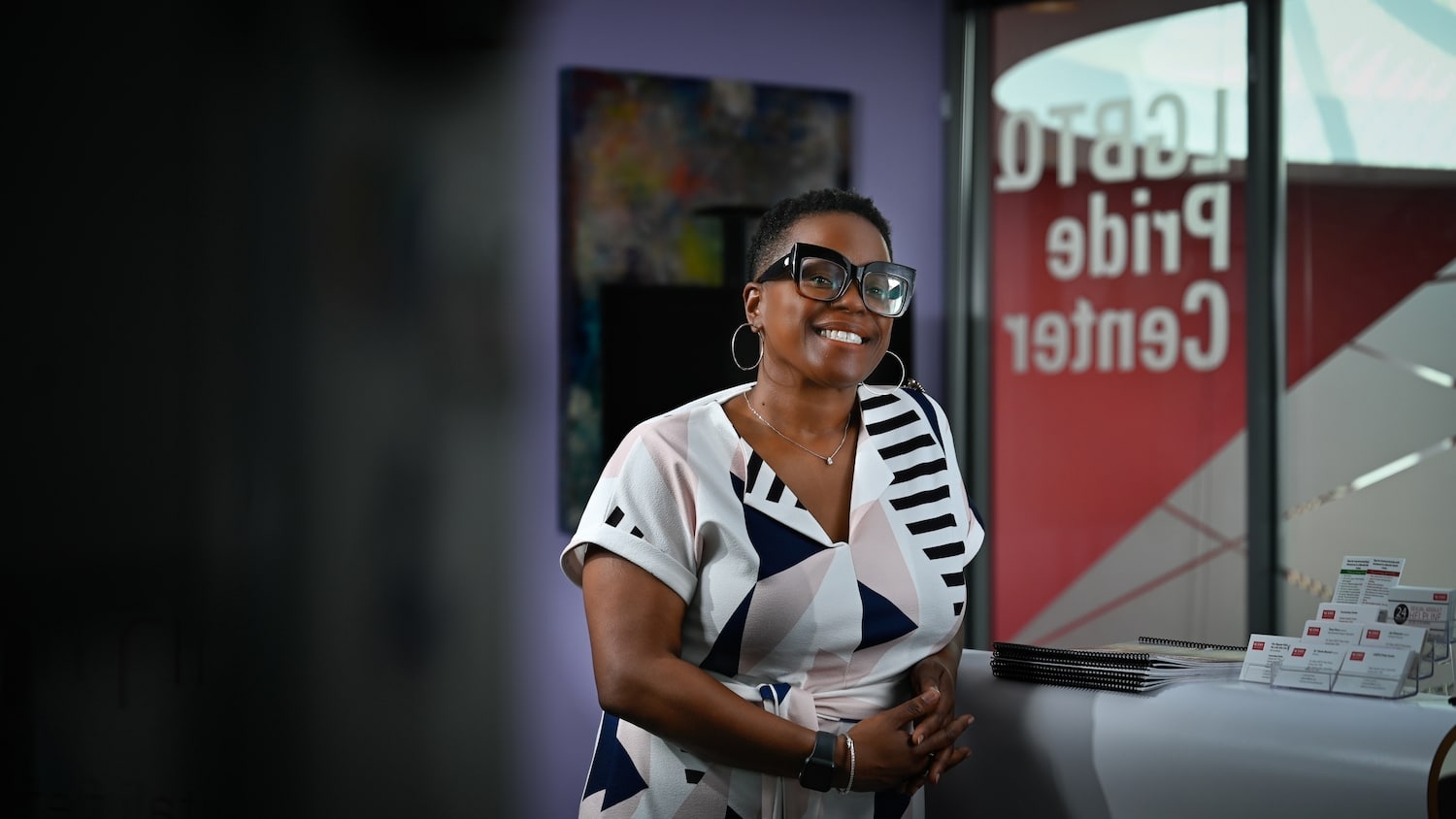 Director of the LGBTQ Pride Center, Charla Blumell, leans on desk and smiles in front the Pride Center's etched glass.